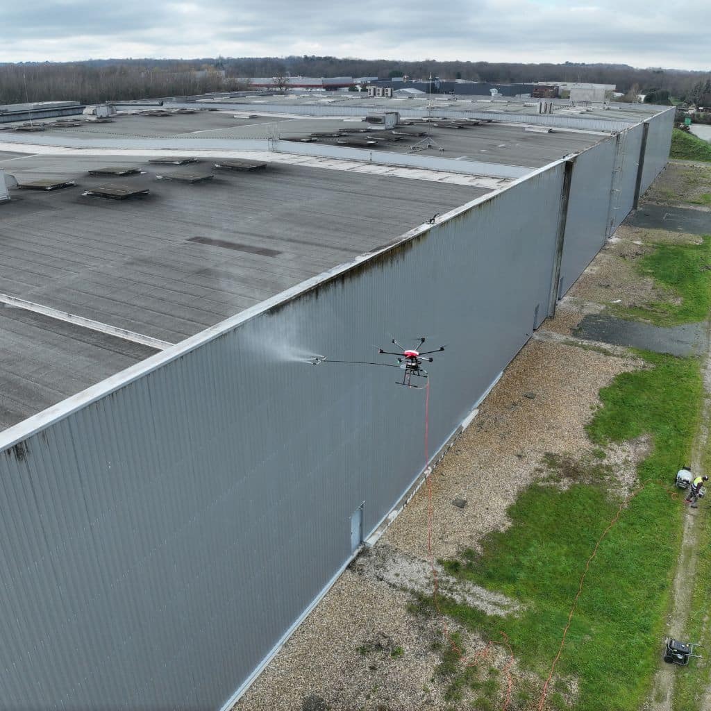 Drone professionnel pulvérisant un agent nettoyant sur le bardage métallique d'un entrepôt logistique de grande hauteur.